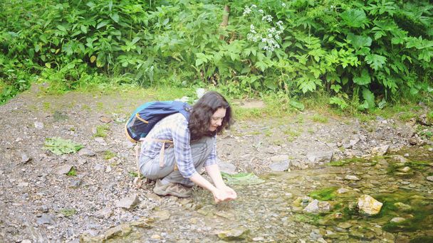 Podróżowanie kobieta mycia jej twarzy i ręce z wodą z strumienia w lesie. Dziewczyna siedzi z plecakiem - Zdjęcie, obraz