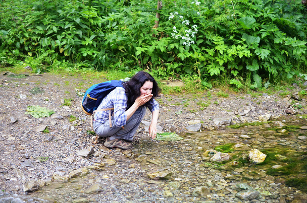 Мандрівна жінка миє обличчя і руки водою з струмка в лісі. Дівчина сидить з рюкзаком
 - Фото, зображення