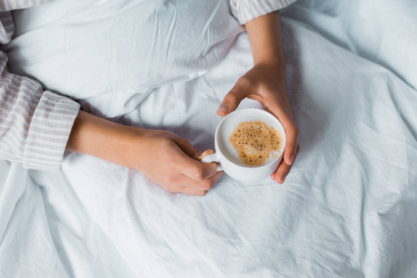 cropped view of woman holding cup of cappuccino in bed  - Photo, Image