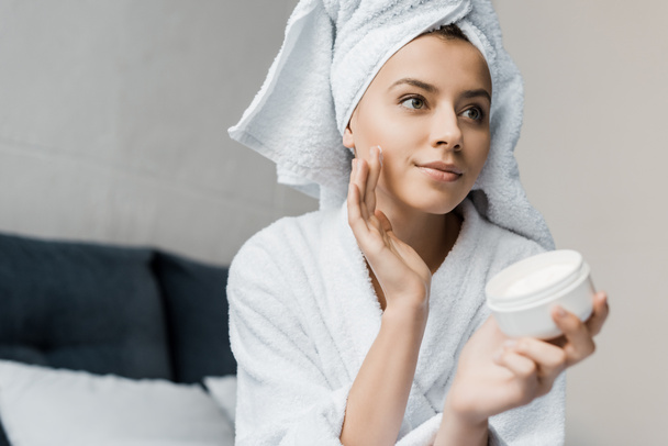 attractive young woman in bathrobe and towel on head applying moisturizing cream on face - Fotó, kép