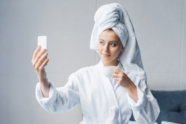 beautiful girl in bathrobe and towel taking selfie with cup of coffee in the morning - Фото, зображення