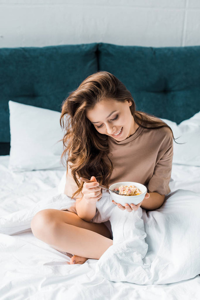 Szczęśliwa dziewczyna jedzenia płatków owsianych na śniadanie, siedząc na łóżku - Zdjęcie, obraz