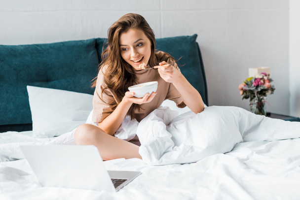 smiling girl eating oatmeal and watching something on laptop while sitting on bed  - Foto, Imagem