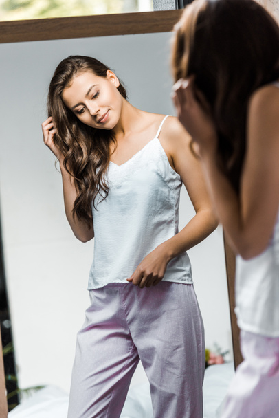 happy brunette girl in pajamas looking at her reflection in mirror - Фото, изображение