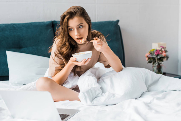 shocked woman eating oatmeal for breakfast and watching something on laptop while sitting on bed - Фото, изображение