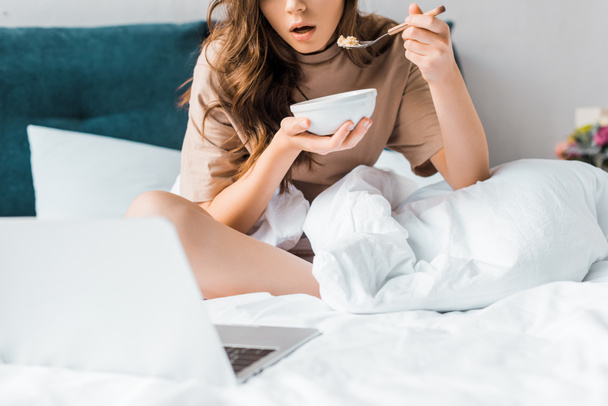 cropped view of shocked girl eating her breakfast and watching something on laptop while sitting on bed - Фото, зображення