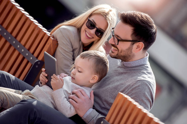 Happy father, mother and son using digital tablet while sitting on the bench in the city. - 写真・画像