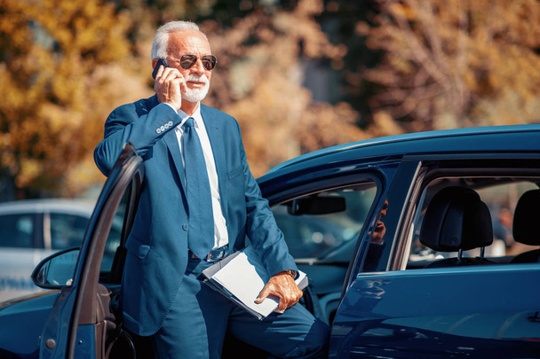 Viaggio d'affari. Fiducioso uomo d'affari senior che utilizza lo smart phone di fronte all'auto
. - Foto, immagini