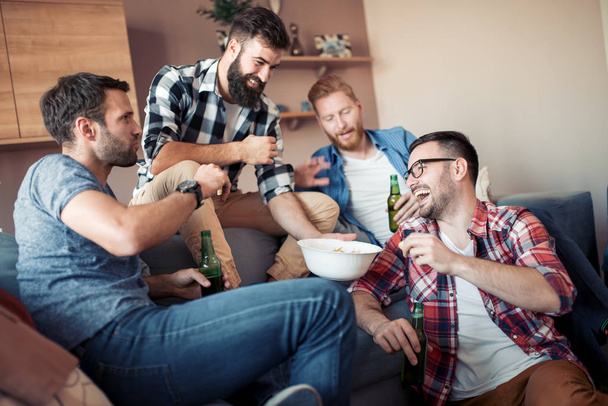 Счастливые молодые парни отдыхают с бутылками напитков, едят попкорн, говорят о футболе и улыбаются.
. - Фото, изображение