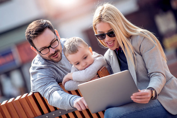 Familia feliz y tecnología digital.Padres jóvenes y niños pequeños usando la computadora portátil mientras están sentados en el parque de la ciudad
. - Foto, Imagen