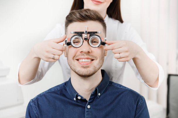 El hombre se sienta y sonríe en un marco de hierro para la selección de lentes de los ojos de recepción oftalmólogo
. - Foto, Imagen