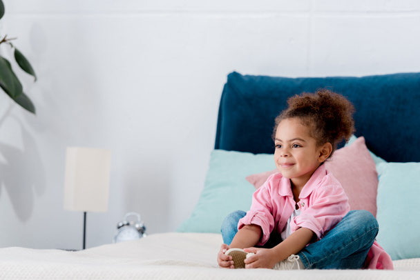 Smiling african american kid siting on the bed clasping her legs - Foto, Imagen