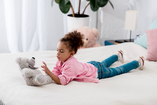 Suloinen kihara afrikkalainen amerikkalainen lapsi makaa sängyllä ja leikkii lelukarhullaan.
 - Valokuva, kuva