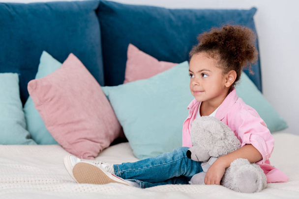 Cute african american child seatting on the bed with her toy bear - Fotó, kép