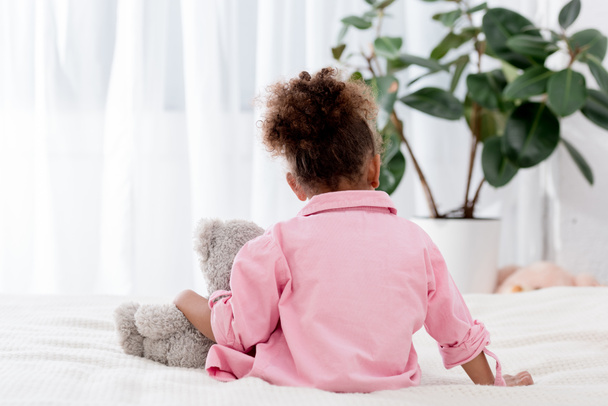 Back view of curly african american child seatting on the bed with her toy bear - Foto, Imagen