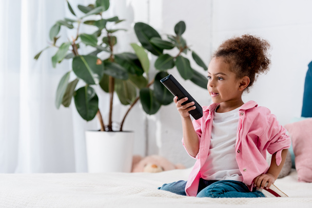 Adorable African american child sitting on the bed and and switching channels on tv - Photo, Image