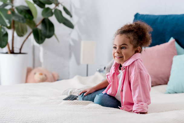 Sonriente niño afroamericano sentado en la cama y viendo el programa de televisión
 - Foto, Imagen
