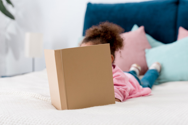 Niño afroamericano rizado acostado en la cama y leyendo libro
 - Foto, Imagen