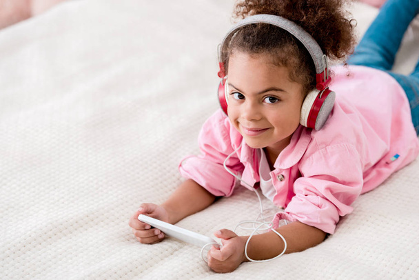 smiling african american child lying on the bed and listening music on her smartphone in headphones - Foto, Bild
