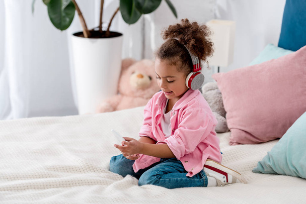 στοχαστικός Αφρικανικός Αμερικανός παιδί κάθεται στο κρεβάτι με ακουστικά και με smartphone  - Φωτογραφία, εικόνα