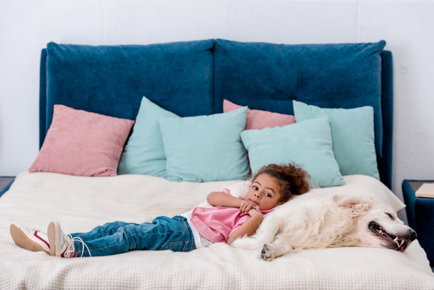 Roztomilý africké americké dítě v růžové sako, ležel na posteli a naklonil se, pes - Fotografie, Obrázek