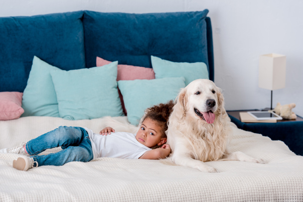 entzückendes kleines afrikanisch-amerikanisches Kind liegt auf dem Bett und lehnt sich an einen glücklichen Hund - Foto, Bild