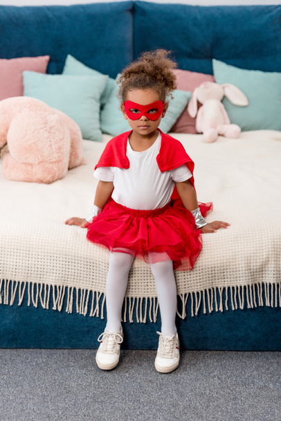 Lindo niño afroamericano en traje de superhéroe rojo y máscara sentado en la cama
 - Foto, imagen