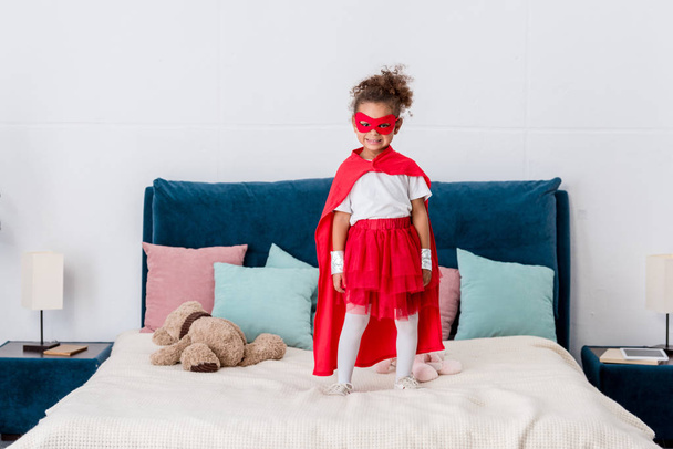 Sorrindo pequena criança afro-americana em traje de super-herói em pé na cama com almofadas coloridas
 - Foto, Imagem