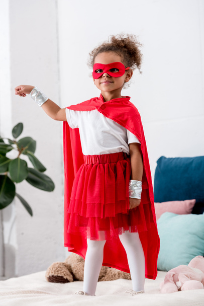 Cute little african american kid in red superhero costume gesturing while standing on bed - Fotó, kép