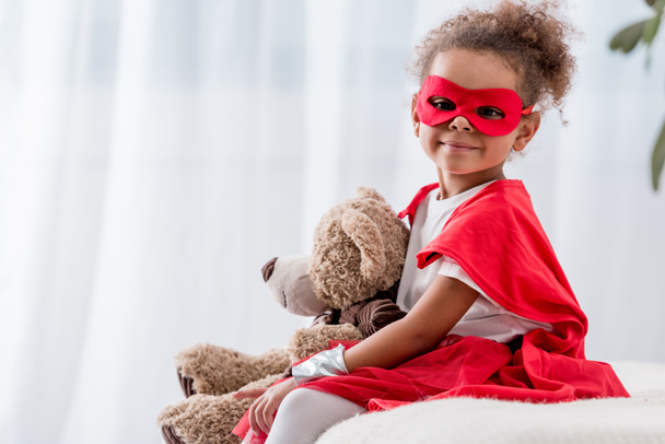 Sevimli küçük Afro-Amerikan çocuk süper kahraman kostümü ve oyuncak ayı ile maske - Fotoğraf, Görsel