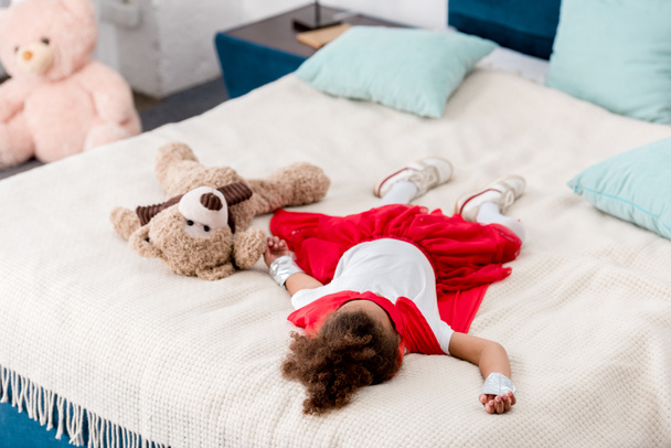 kindje in rode superheld kostuum met teddybeer liggend op bed - Foto, afbeelding