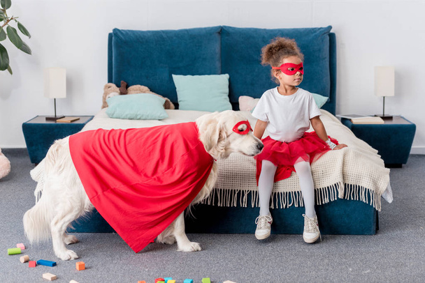 Cute little african american kid with dog in superhero costumes in bedroom - Photo, image