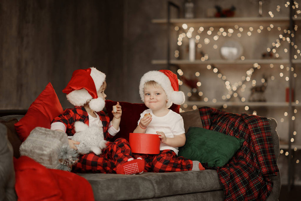 Little cute kids, brother and sister, wearing Santa Claus hats, are sitting on a big easy chair in the living room and are looking at Christmas presents in boxes.  - Photo, Image