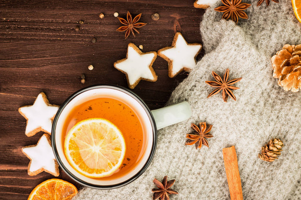 Winter theme. Christmas tea with spices, cup of tea with orange, cinnamon, anise, cookies in a shape of star, pepper, fir cones and gray scarf on wooden background. Flat lay, View from above. Copy space for text. - Photo, Image