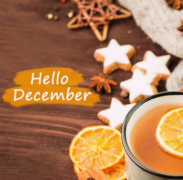 Winter theme. Cup of hot tea with spices, orange, cinnamon, anise, cookies in a shape of star, pepper and gray scarf on wooden background with text hello december. View from above. - Φωτογραφία, εικόνα