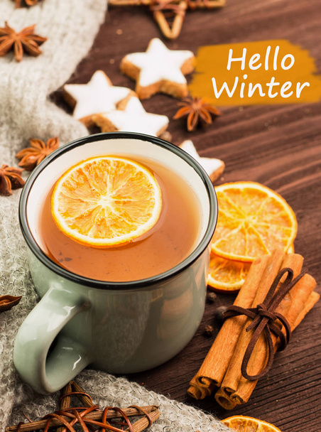 Winter theme. Cup of hot tea with spices, orange, cinnamon, anise, cookies in a shape of star,  pepper and gray scarf on wooden background with text hello winter. View from above, - Photo, Image