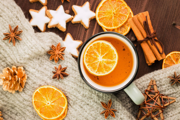 Winter theme. Christmas tea with spices, cup of tea with orange, cinnamon, anise, cookies in a shape of star, pepper, fir cones and gray scarf on wooden background. Flat lay, View from above. - Photo, Image