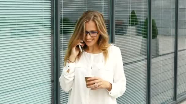 4K. Atractiva mujer de negocios, se acerca a la oficina, bebe café, sonríe y habla por teléfono inteligente
 - Imágenes, Vídeo