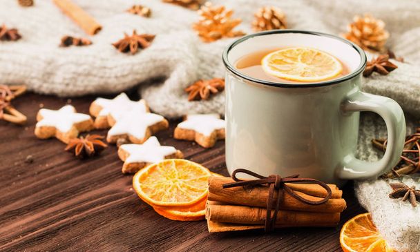 Winter and New Year theme. Christmas tea with spices, cup of tea with orange, cinnamon, anise, cookies in a shape of star, fir cones, pepper and gray scarf on wooden background. Copy space for text. - Photo, Image