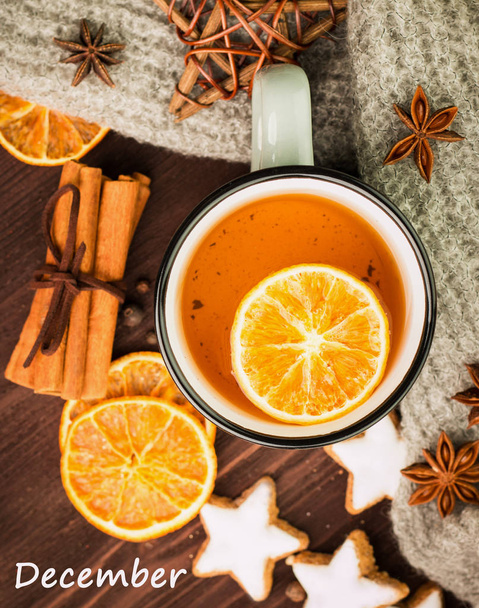 Winter theme. Hot tea with spices, orange, cinnamon, anise, cookies in a shape of star, pepper and gray scarf on wooden background. Flat lay, View from above with text december. - Photo, Image