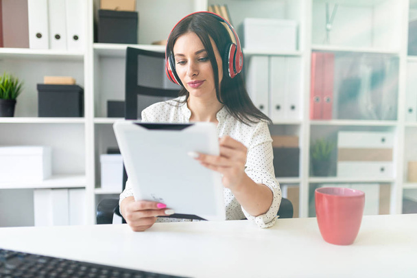 Mladá dívka se sluchátky na uších sedí v kanceláři na stole počítač a drží tablet. - Fotografie, Obrázek
