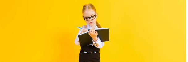 Menina com cabelo vermelho em um fundo amarelo. Uma garota encantadora em óculos transparentes escreve uma caneta em seu caderno
. - Foto, Imagem