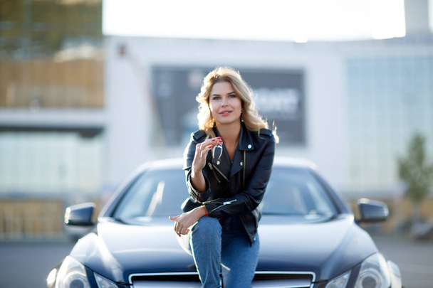 Image of smiling blonde with keys sitting on hood of black car - Zdjęcie, obraz