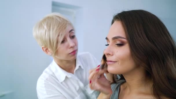 closeup blonde makeup artist doing professional makeup for brunette cute woman at beauty salon - Video
