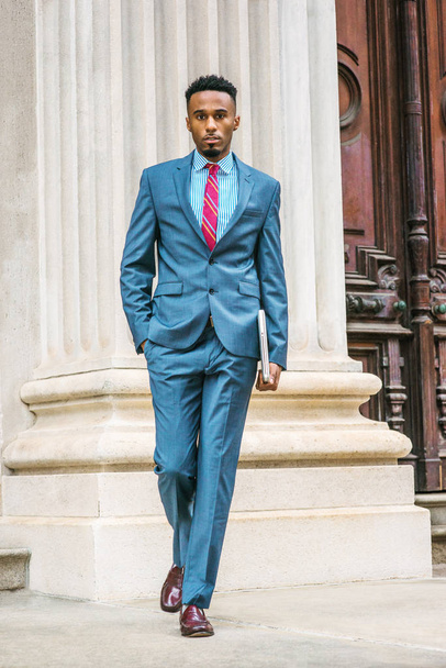 Fiatal afro-amerikai üzletember, a szakáll működő New York-i, ég-kék öltöny, lila, piros mintás nyakkendő, sötét lila bőr cipő viselése, gazdaság laptop számítógép, kisétált az iroda ajtaja út. - Fotó, kép