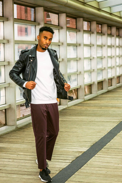 Young Man Casual Fashion in New York. Young African American Guy with beard, wearing black leather jacket, white shirt, black pants, sneakers, standing on walk way with glass wall and wooden floor. - 写真・画像