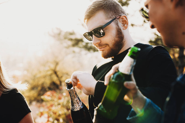 Young bearded guy in sunglasses opening bottle of champagne while celebrating with friends in countryside - Fotografie, Obrázek
