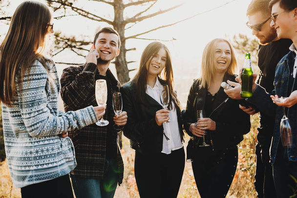 Веселые молодые друзья, улыбающиеся и пьющие пиво и шампанское, стоя в прекрасной сельской местности вместе
 - Фото, изображение