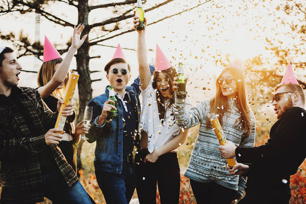 Ryhmä nuoria, jotka nostavat pulloja olutta ja räjähtäviä juhlakeksejä värikkäillä konfetteilla viettäen joulua luonnossa.
 - Valokuva, kuva