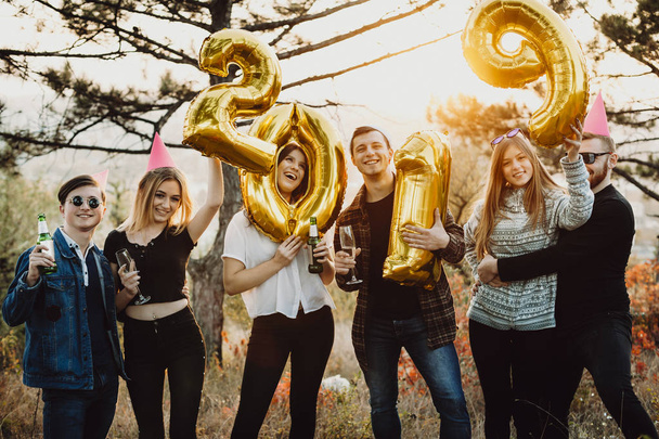 Группа молодых людей в повседневных нарядах держит бутылки пива и золотые воздушные шары, формирующие 2019 письменной форме во время празднования Нового года в природе
 - Фото, изображение